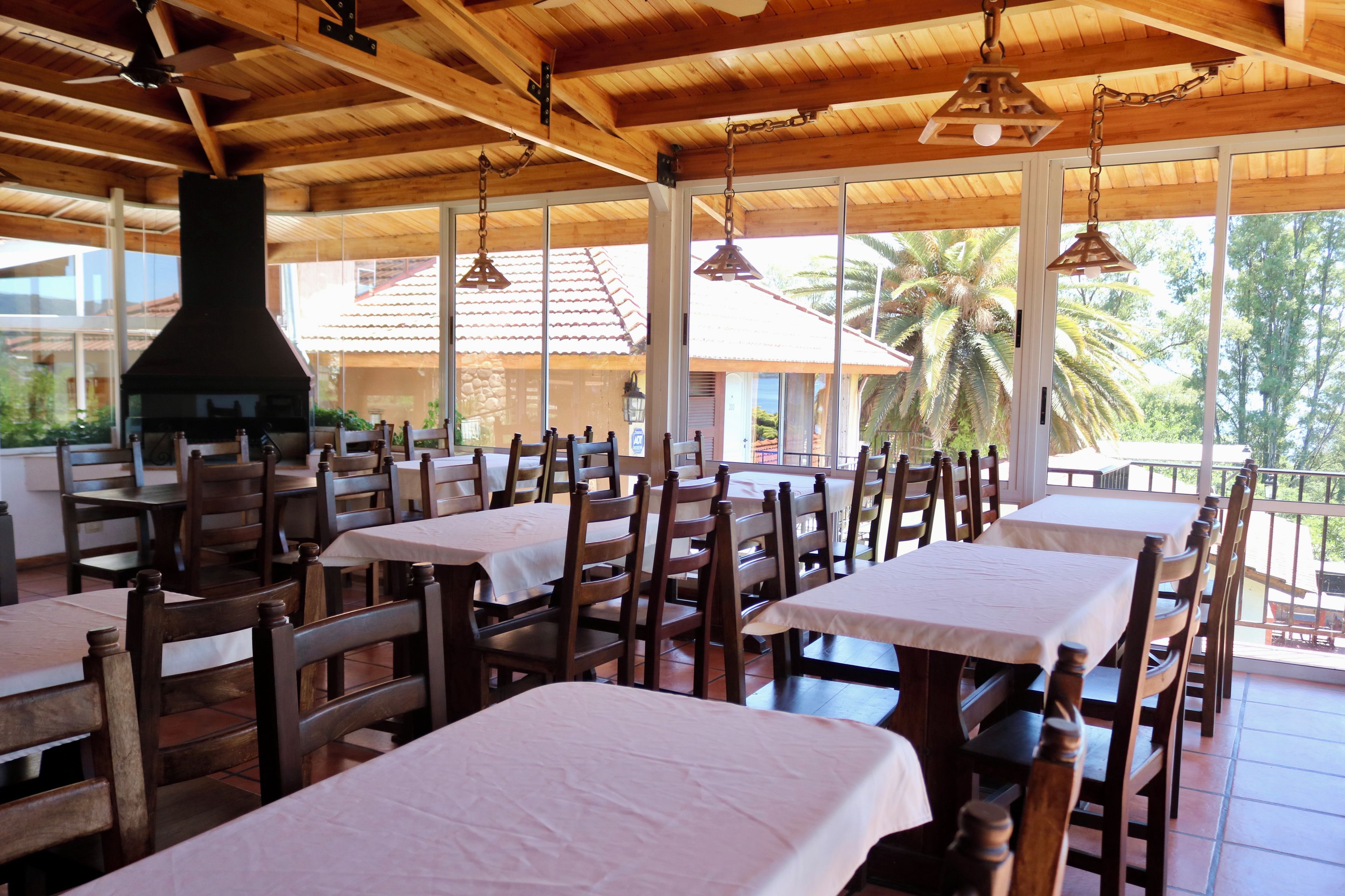 Un restaurant avec des tables et des chaises en bois, une grande cheminée et de grandes fenêtres donnant sur des palmiers.
