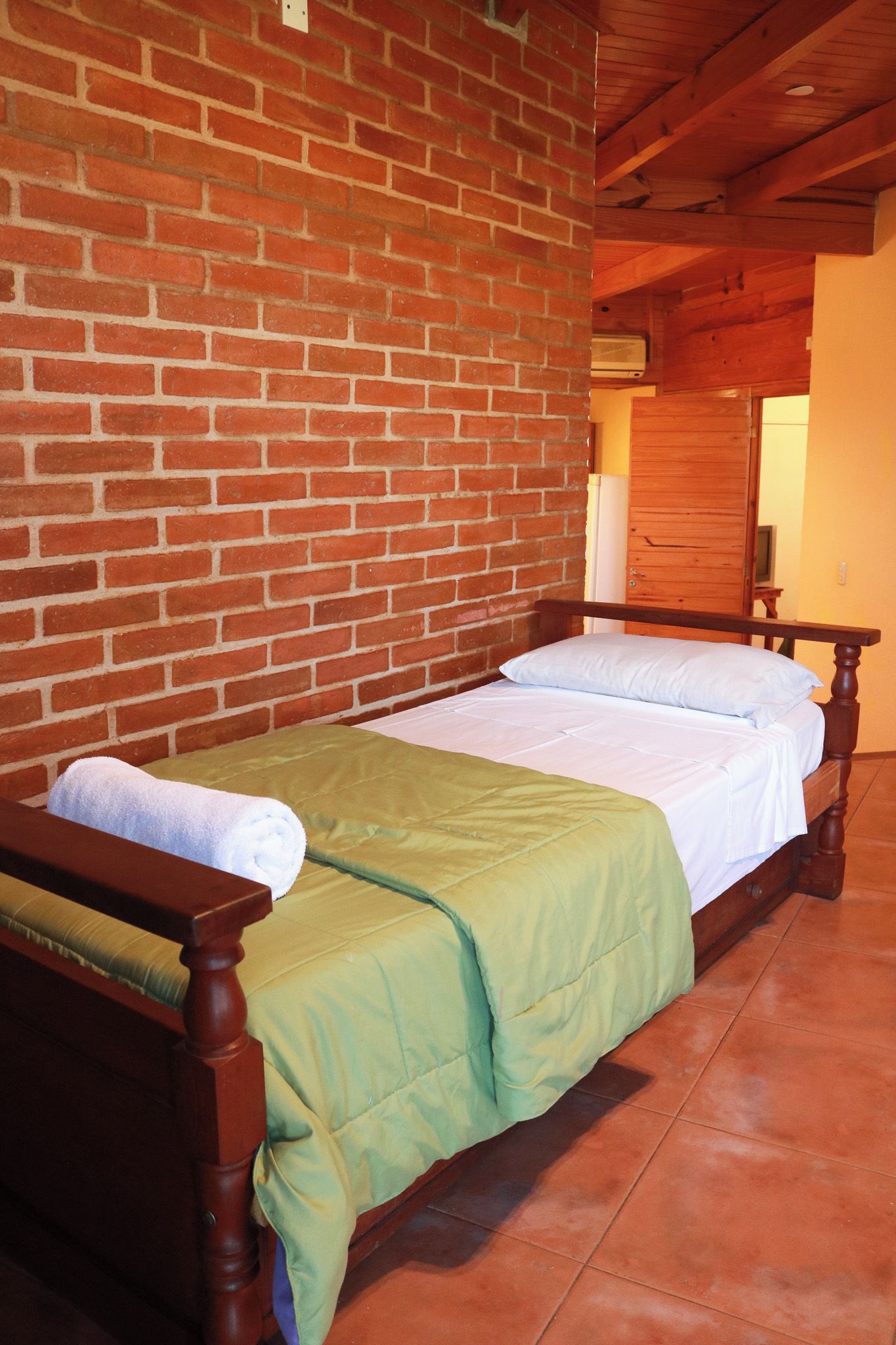 Una cama individual con estructura de madera y edredón verde en una habitación con paredes de ladrillo y vigas de madera.