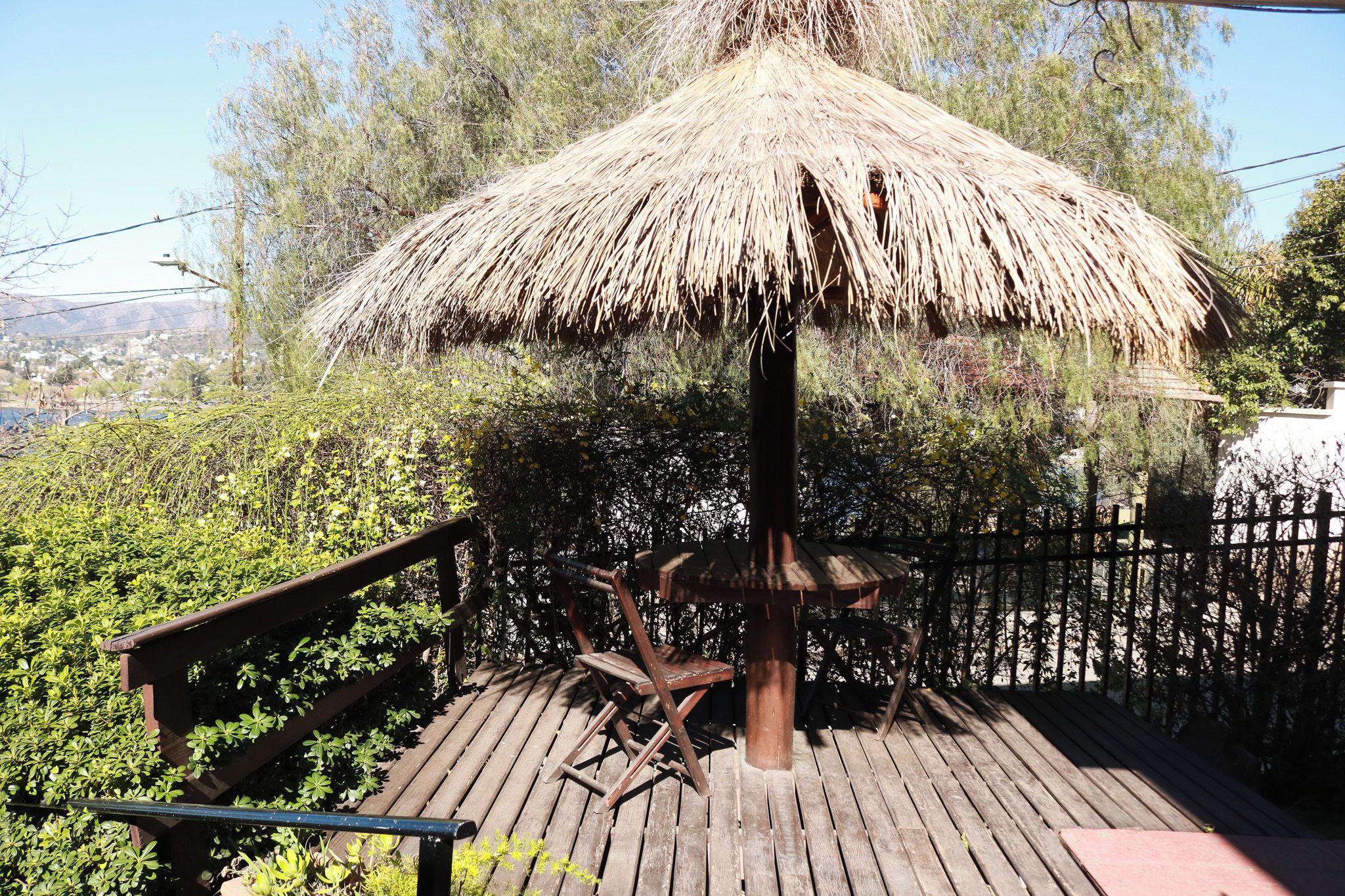 Una terraza de madera con estructura de techo de paja, rodeada de follaje y una valla.