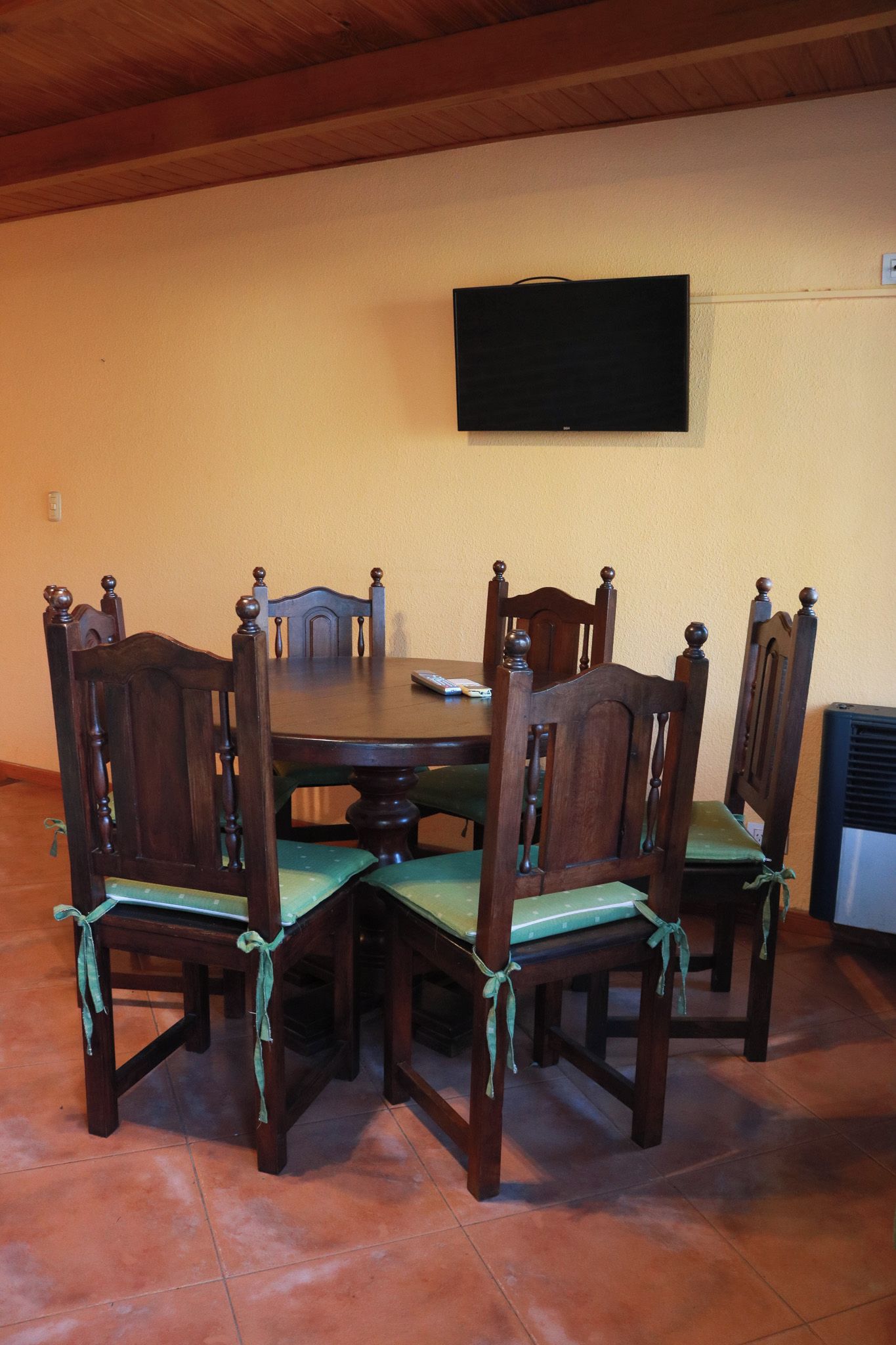A dining area with a wooden table and chairs, a flat-screen TV mounted on the wall, and a tiled floor.