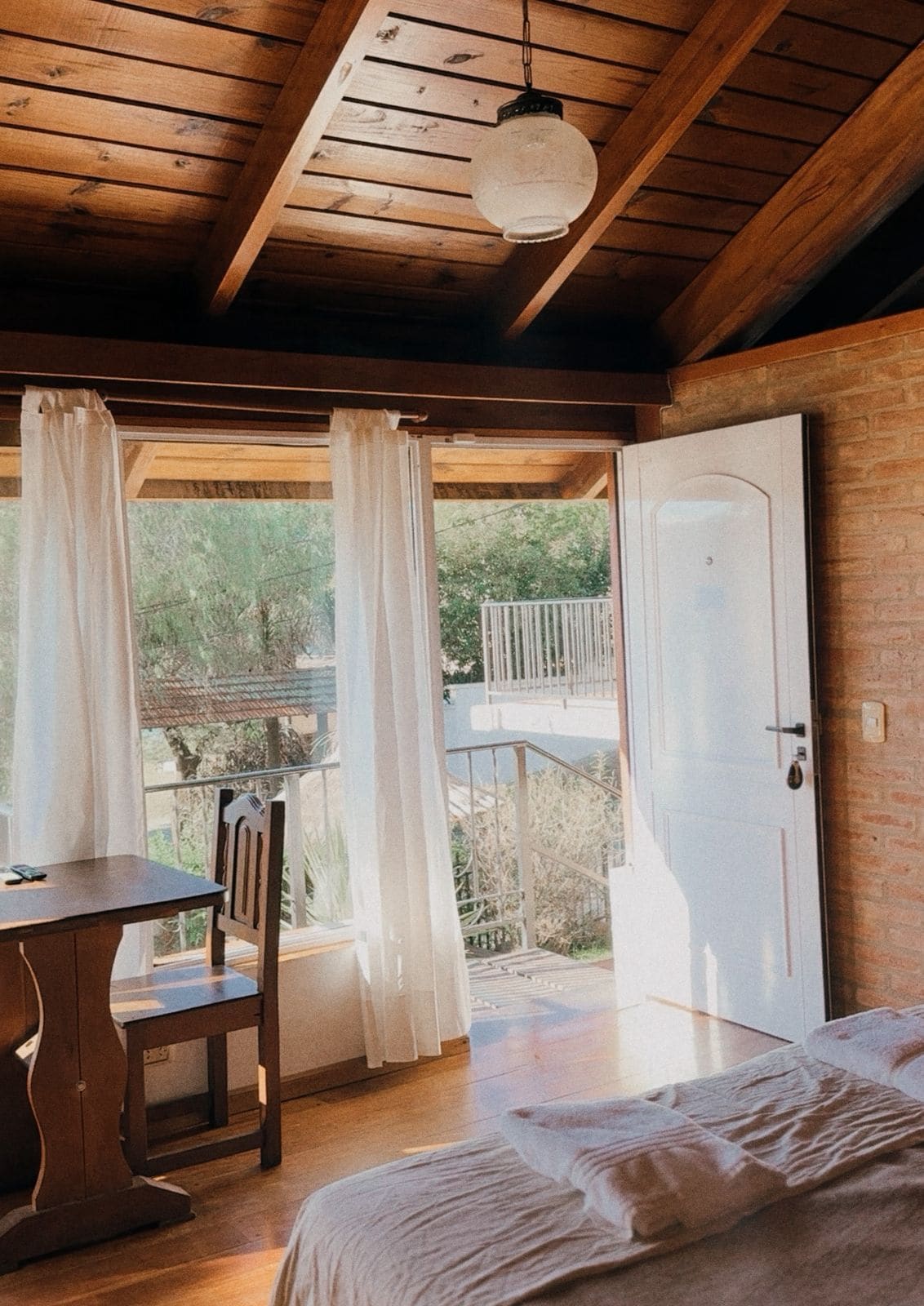 Una habitación con techo de madera, una cama, una mesa con una silla y vistas a un balcón exterior.