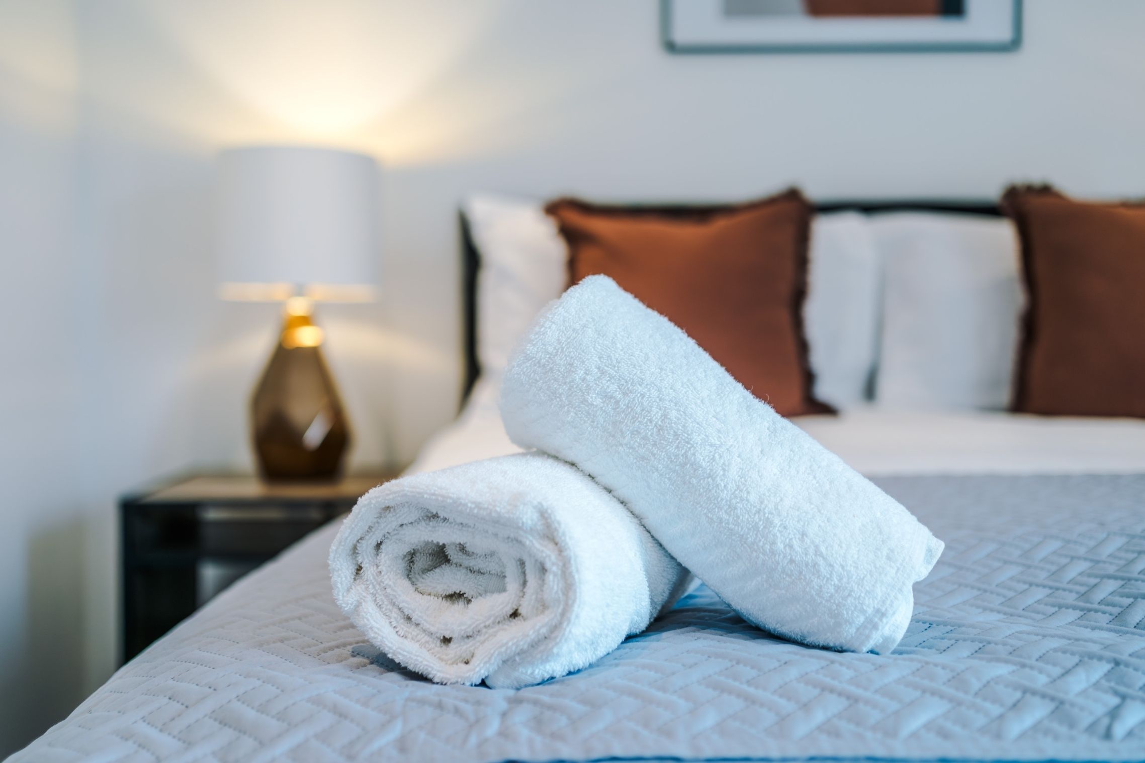 A neatly made bed with a rolled white towel, a bedside lamp, and brown pillows.