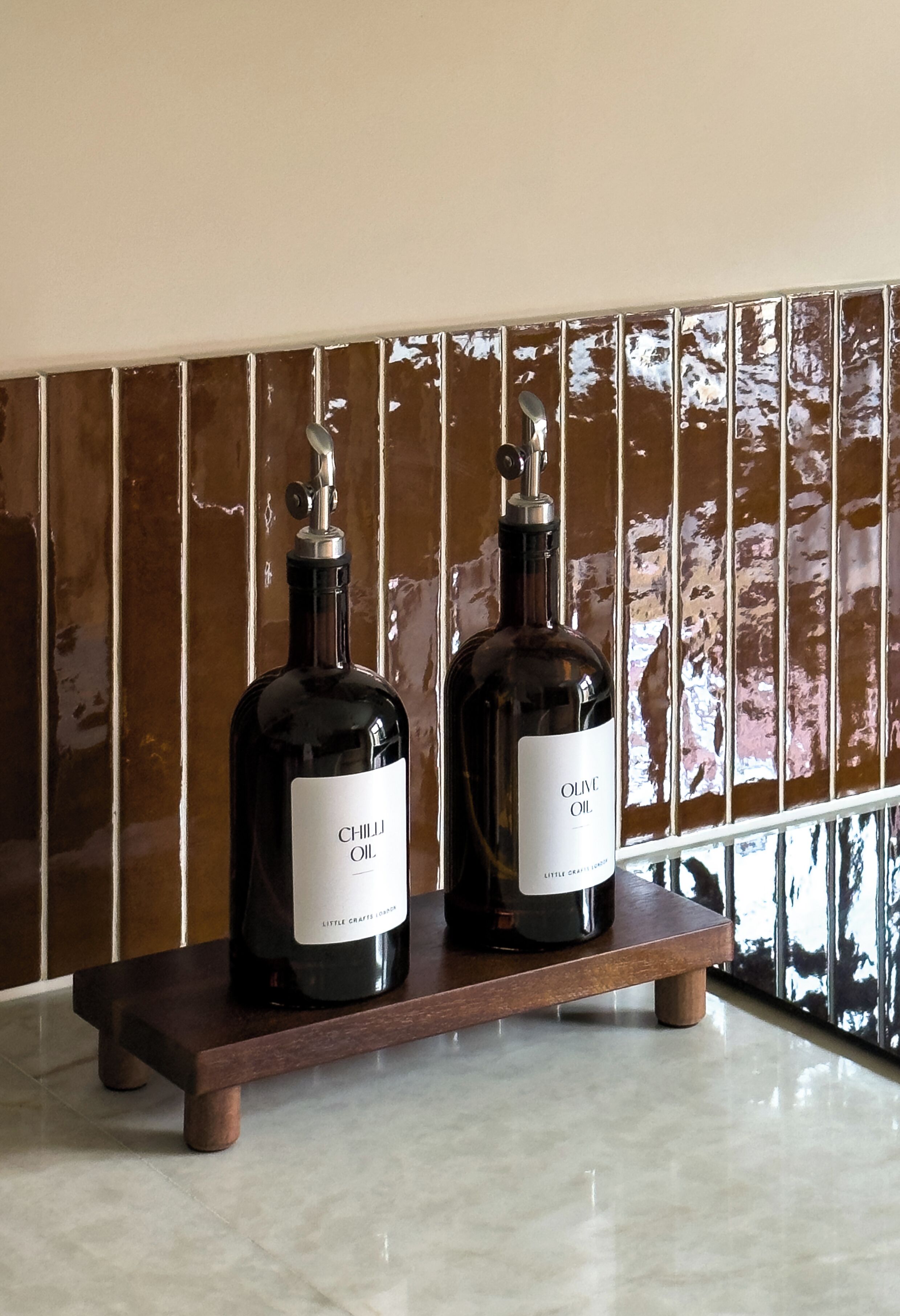 Deux bouteilles d’huile d’olive sont posées sur un présentoir en bois, devant un mur recouvert de carreaux.