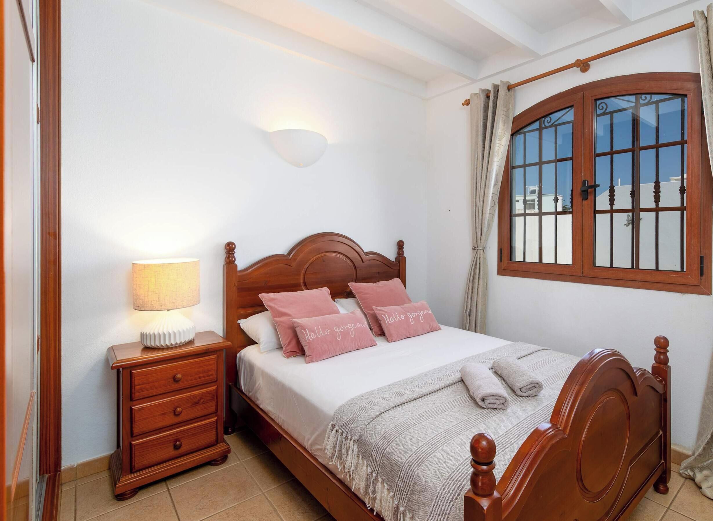 A bedroom with a wooden bed, bedside table with a lamp, a window with curtains, and a wall-mounted light fixture.