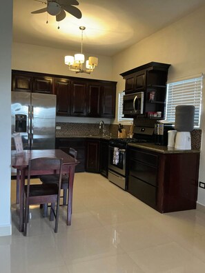 A kitchen with dark wooden cabinets, stainless steel appliances, a dining table with chairs, and a ceiling fan. - Garden City Suite | Modern 2BR Apartment (Belmopan)