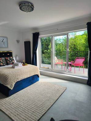 A bedroom with a large bed, a nightstand, a chandelier, a window with a view of greenery, and a balcony with chairs. - House near Windsor with river view (Wraysbury)
