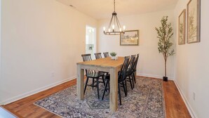 A dining area with a wooden table and chairs, a chandelier, a patterned rug, and a potted plant. - Pool Home w/ King Beds, 4 TVs & Large Backyard (Valdosta)