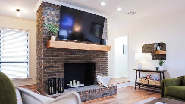 A living room with a brick fireplace, a flat-screen TV mounted above it, a wooden coffee table, and a green armchair. - Pool Home w/ King Beds, 4 TVs & Large Backyard (Valdosta)