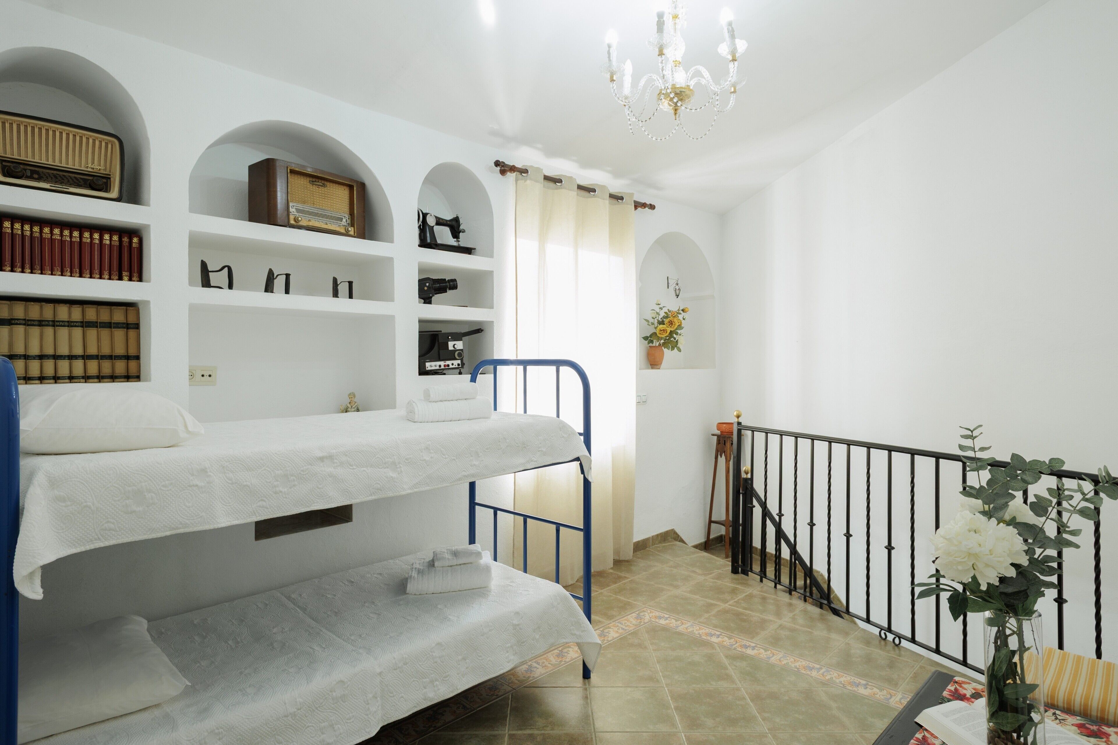 A room with bunk beds, a bookshelf with books and a radio, a chandelier, and a staircase with a railing.