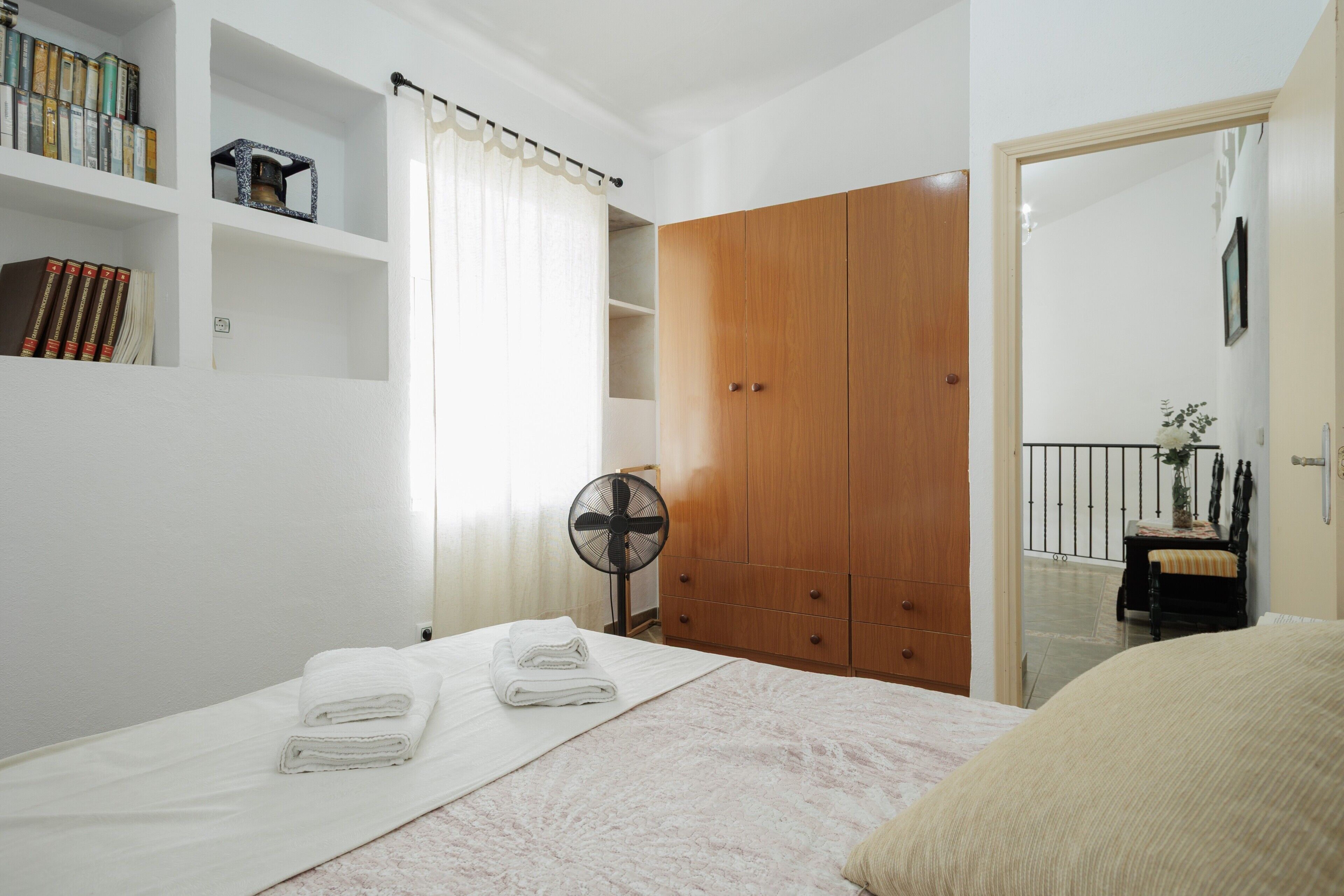Un dormitorio con cama, un armario de madera, un ventilador y una estantería con libros.