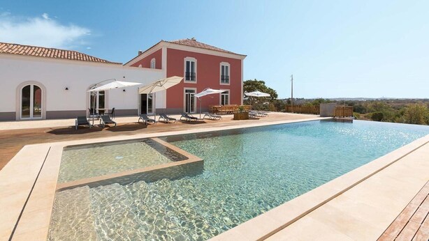 Piscina com espreguiçadeiras e guarda-sóis, um edifício vermelho com janelas em arco e céu claro.
