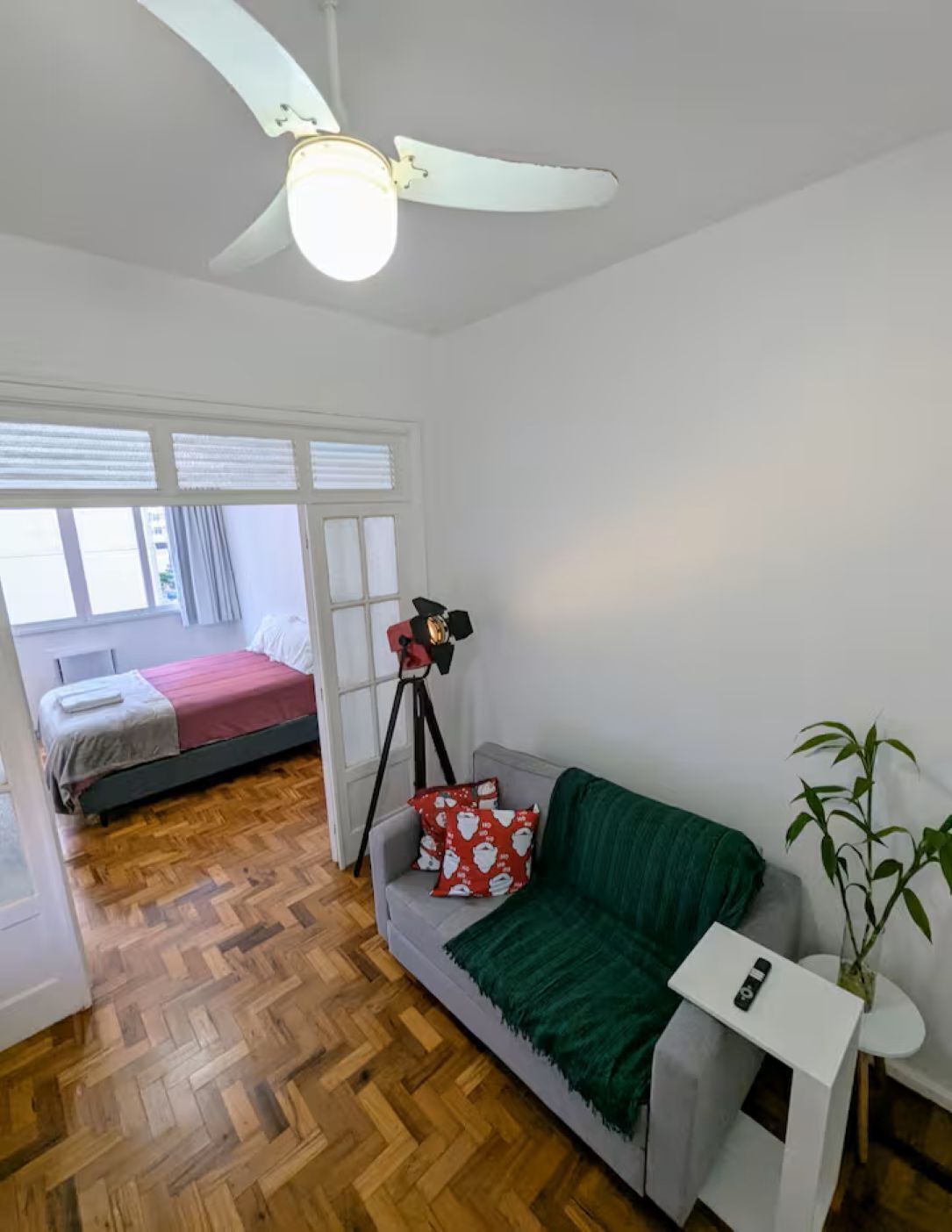 Een kamer met een bed, een slaapbank, een plafondventilator, een statief en een potplant.