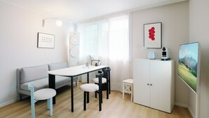 Une pièce moderne et bien éclairée, avec une table blanche, des chaises, un canapé, un meuble, un téléviseur et une fenêtre ornée de rideaux légers.