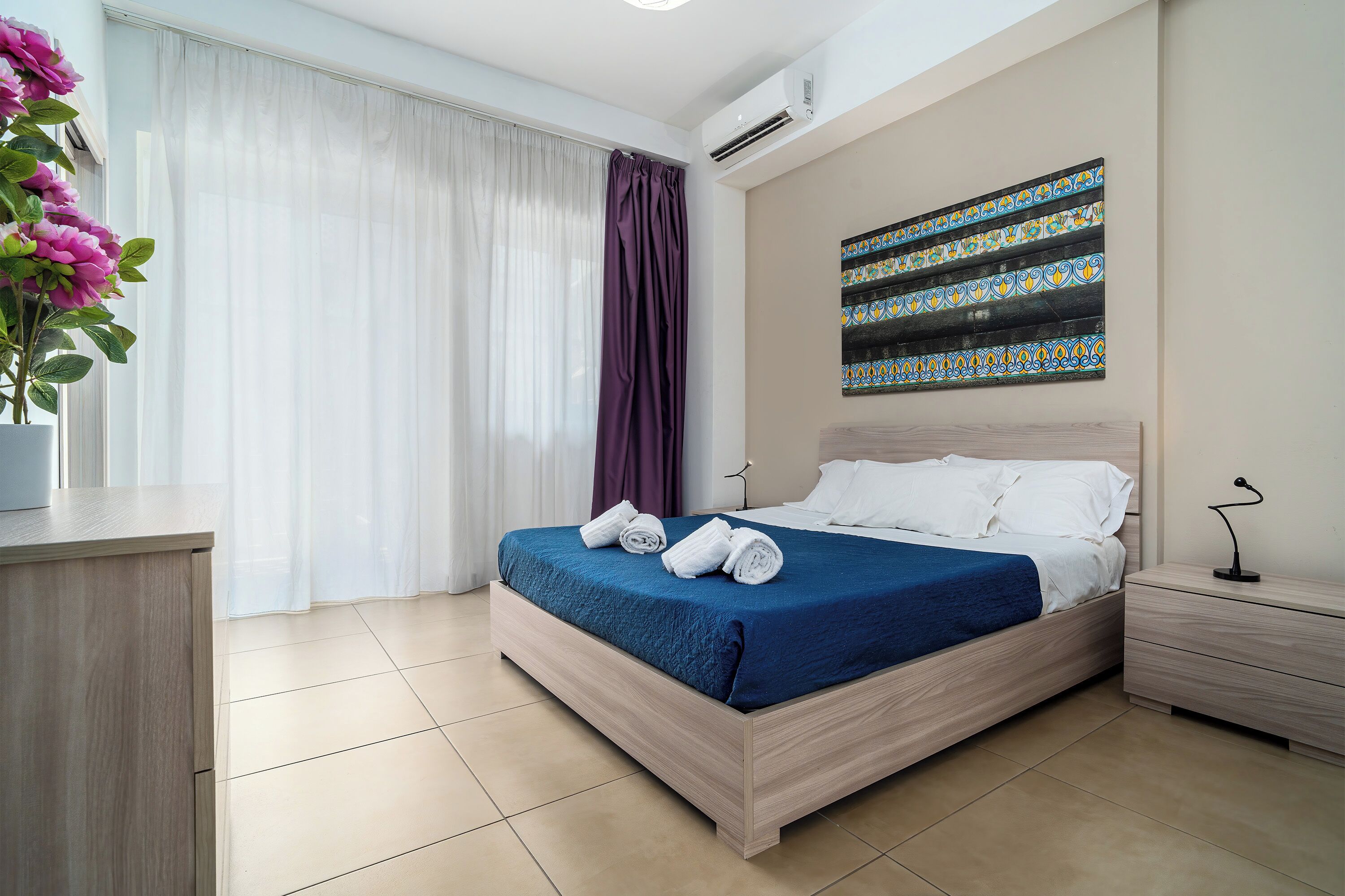 Ein Hotelzimmer mit Bett, Nachttisch, Schreibtisch, einer Zimmerpflanze und einem Fenster mit Vorhängen.