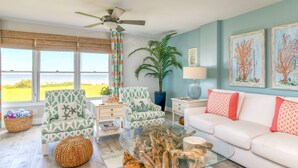 A living room with a view of the ocean, a ceiling fan, a large plant, and a white sofa with red cushions. - Sand Dollar 103 (St. Augustine)