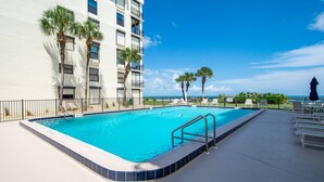 A swimming pool with a blue border, surrounded by a white building with balconies, palm trees, and a clear sky. - Sand Dollar 103 (St. Augustine)