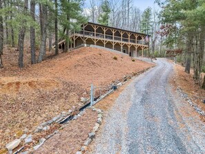 Ein rustikales Lodge mit überdachter Veranda, einem Schotterweg und bewaldetem Hintergrund.