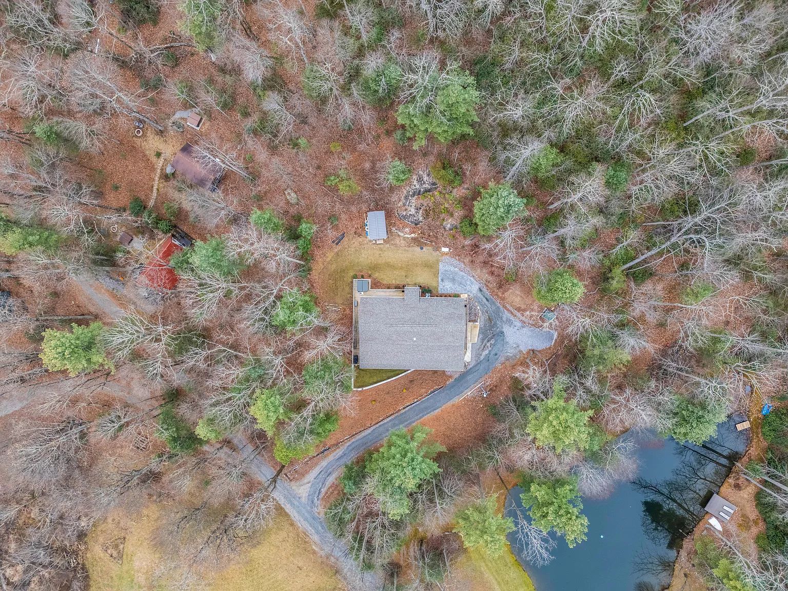 An aerial view of a property with a house, a driveway, a small pond, and surrounding trees.