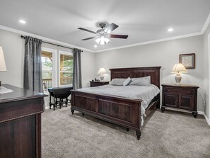 Ein Schlafzimmer mit einem großen Bett, einem Schreibtisch, einem Stuhl, einem Nachttisch, einer Lampe, einem Deckenventilator und einem Fenster mit Vorhängen.