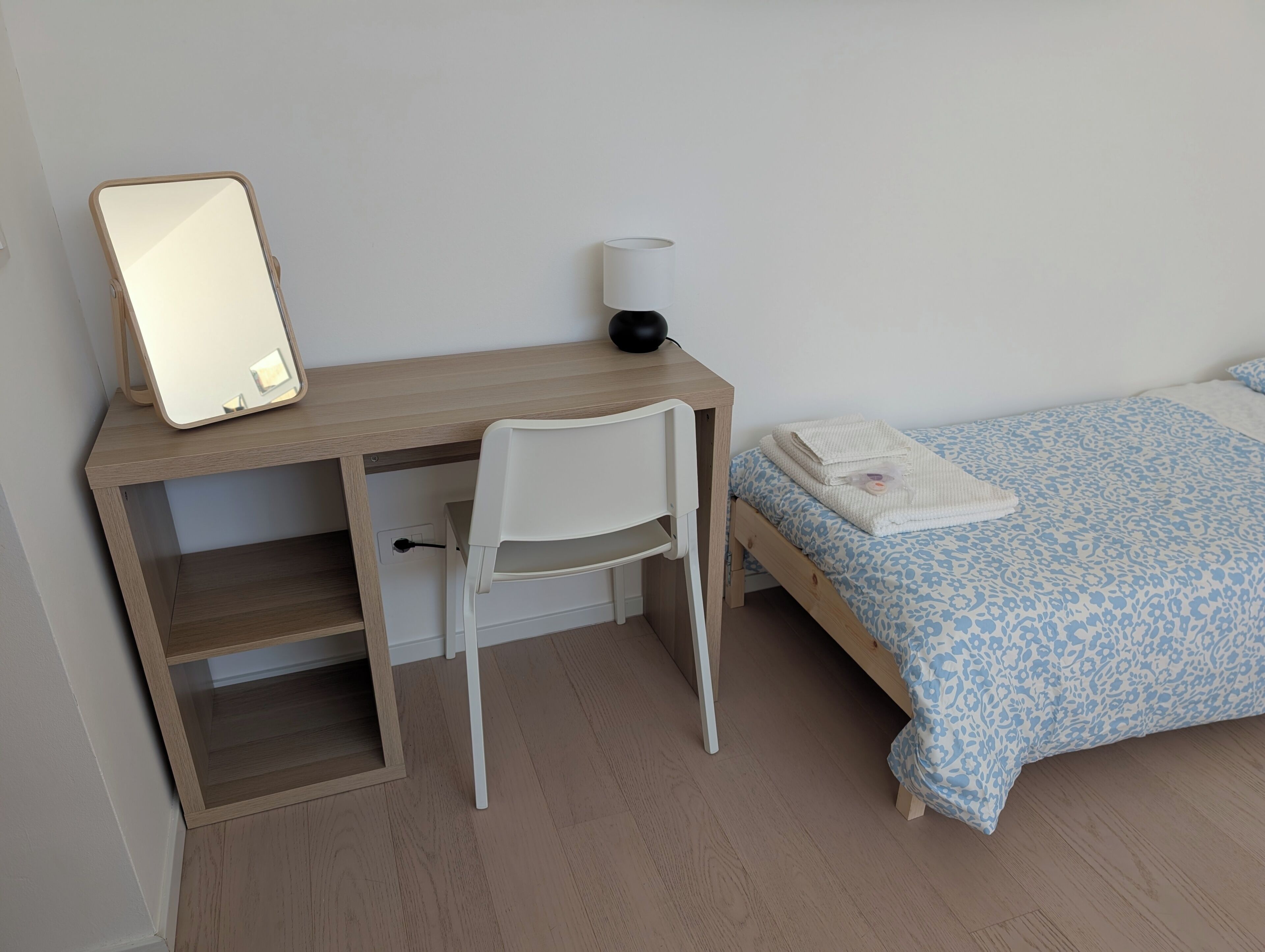 Ein Schlafzimmer mit einem Holztisch, einem weißen Stuhl, einem Bett mit blühendem, blauem Überwurf und einer Nachttischlampe.
