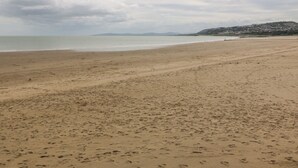 A wide sandy beach with footprints, a calm sea, and a distant hill with buildings. - Beach View Apartment (Colwyn Bay)