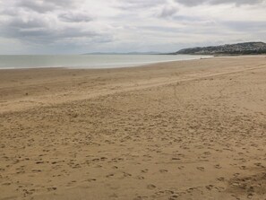 A wide sandy beach with footprints, a calm sea, and a distant hill with buildings. - Beach View Apartment (Colwyn Bay)