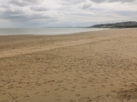 Una playa extensa de arena con huellas, un mar tranquilo y una colina distante con edificios.