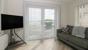 A living room with a television on a stand, a grey sofa with a patterned pillow, and a sliding glass door leading to an outdoor area. - Beach View Apartment (Colwyn Bay)