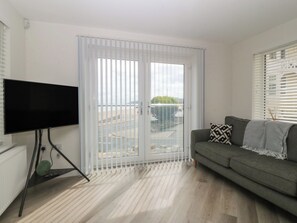 A living room with a television on a stand, a grey sofa with a patterned pillow, and a sliding glass door leading to an outdoor area. - Beach View Apartment (Colwyn Bay)