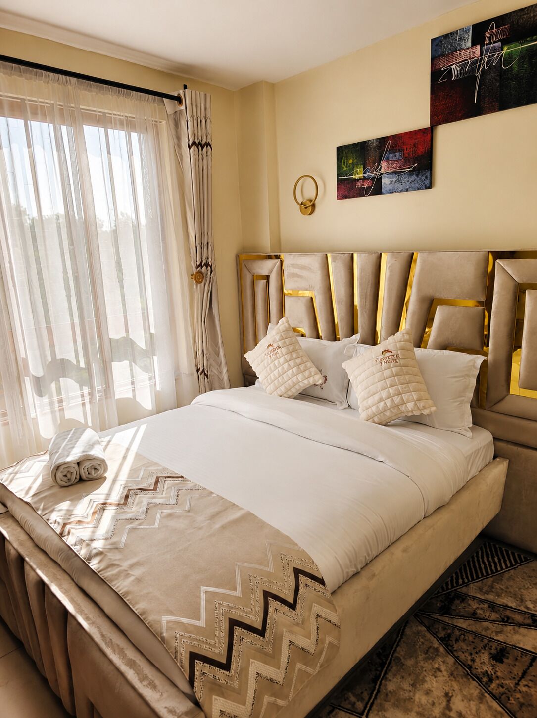A hotel room with a bed, curtains, a wall art piece, and a decorative wall light.