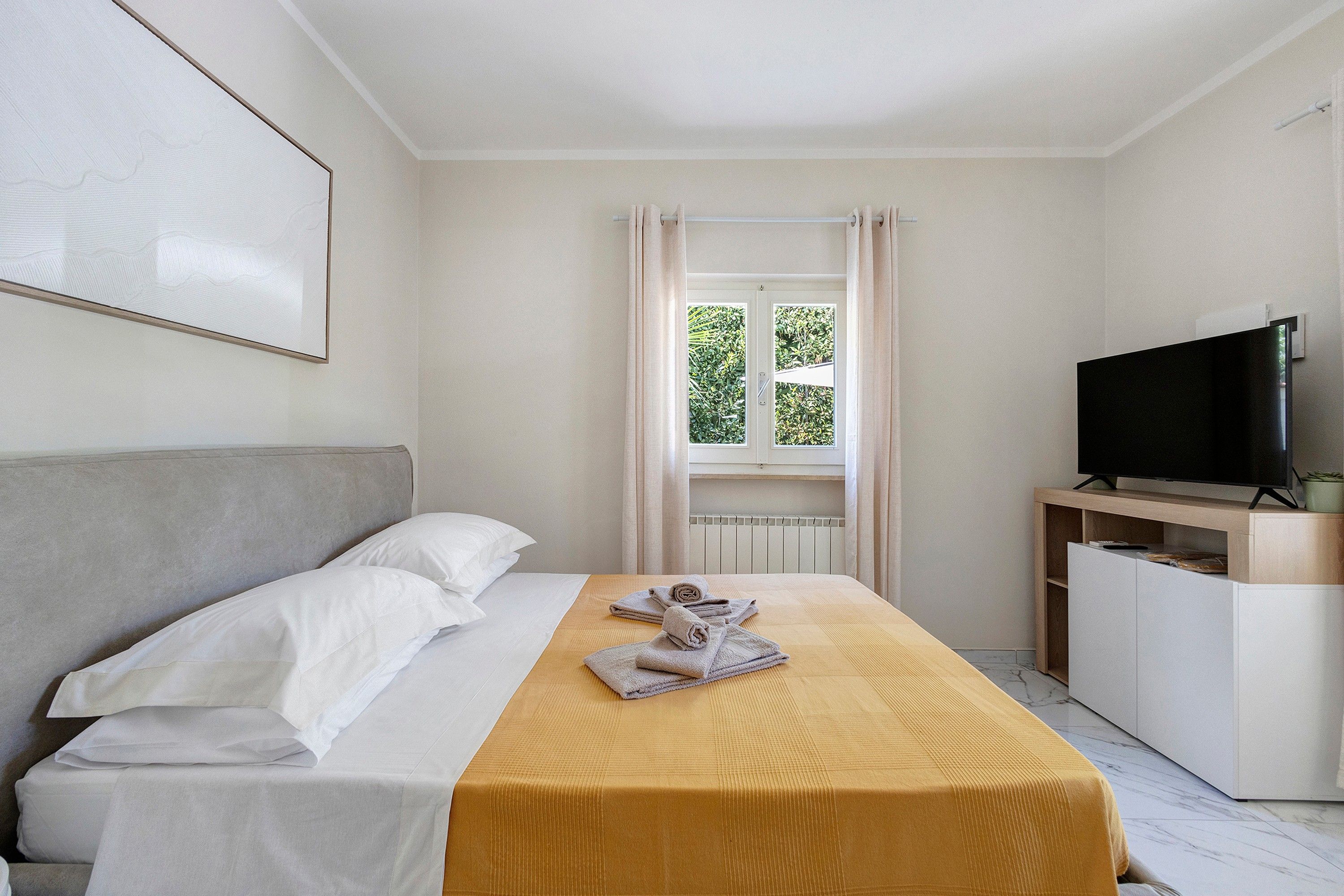 A hotel room with a bed, a television on a stand, a window with curtains, and a radiator.