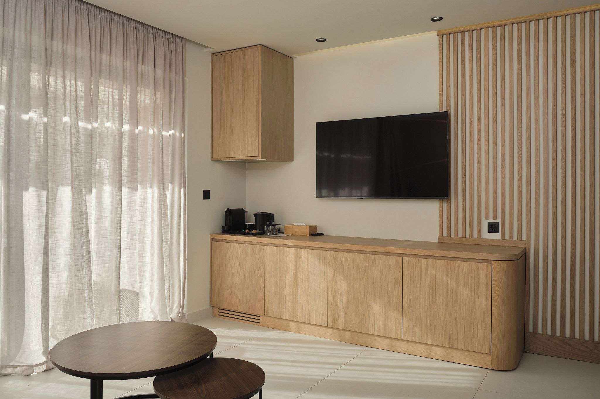 Una habitación moderna con un armario de madera, un televisor de pantalla plana, una mesa redonda y una ventana con cortinas translúcidas.