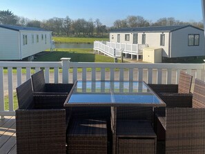 Eine Terrasse mit Korbmöbeln und einem Glastisch, mit Blick auf eine Rasenfläche mit Teich und Fertighäusern.