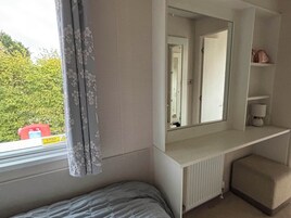Un dormitorio con cama, radiador, espejo, ventana con cortinas y estante con lámpara.