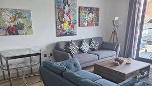Un salon moderne avec un canapé gris, une table basse en verre et une table à manger avec des chaises. Trois grandes œuvres d’art ornent le mur au-dessus du canapé.