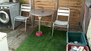 Un pequeño espacio al aire libre con una lavadora, dos sillas de metal, una mesa redonda y un contenedor verde.