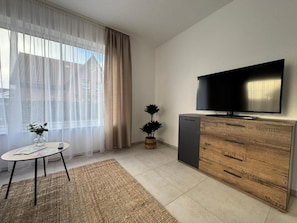 Ein modernes Wohnzimmer mit Flachbildfernseher, einer Holzkommode, einem kleinen Tisch mit einer Vase und transparenten Vorhängen.