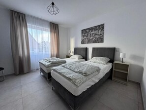 Ein modernes Hotelzimmer mit zwei Betten, einem großen Fenster mit Vorhängen, Blick auf die Stadt und einer geometrischen Leuchte.