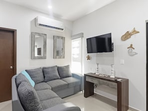 Un salon moderne avec un canapé d’angle gris, un téléviseur à écran plat fixé au mur et une petite table d’appoint en bois.