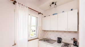 Cocina compacta con armarios blancos, una ventana con cortinas, un lavabo, una estufa y una tetera.