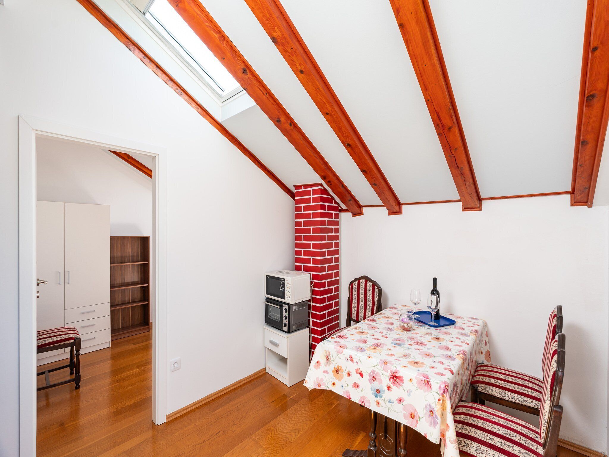 Una habitación con techo inclinado, vigas de madera, pared de ladrillo, una mesa con mantel floral, una silla, microondas y una botella de vino.