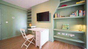 Ein Zimmer mit Schreibtisch, Stuhl, Bücherregal und einem an der Wand befestigten Fernseher.
