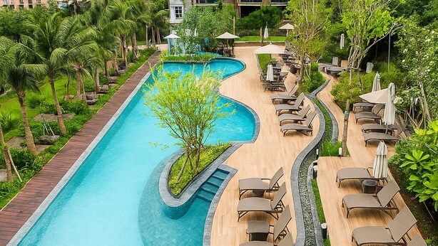 A large, curved swimming pool with lounge chairs and umbrellas, surrounded by lush greenery and a small waterfall feature. - High Rise Luxury Stay Near Beach & Walking Street (ตำบล หนองปรือ)