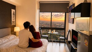 A couple sitting on a bed facing a city view, with a table and chairs in the foreground and a television on a stand. - High Rise Luxury Stay Near Beach & Walking Street (ตำบล หนองปรือ)