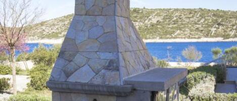 A stone-built outdoor structure with a chimney, overlooking a coastal landscape with a clear view of the sea and hills.