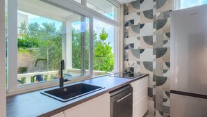 A modern kitchen with a large window, a dishwasher, and a refrigerator. - Secret Garden Apartment Žarka (Postira)