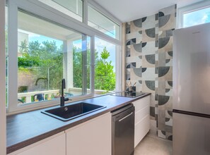 A modern kitchen with a large window, a dishwasher, and a refrigerator. - Secret Garden Apartment Žarka (Postira)