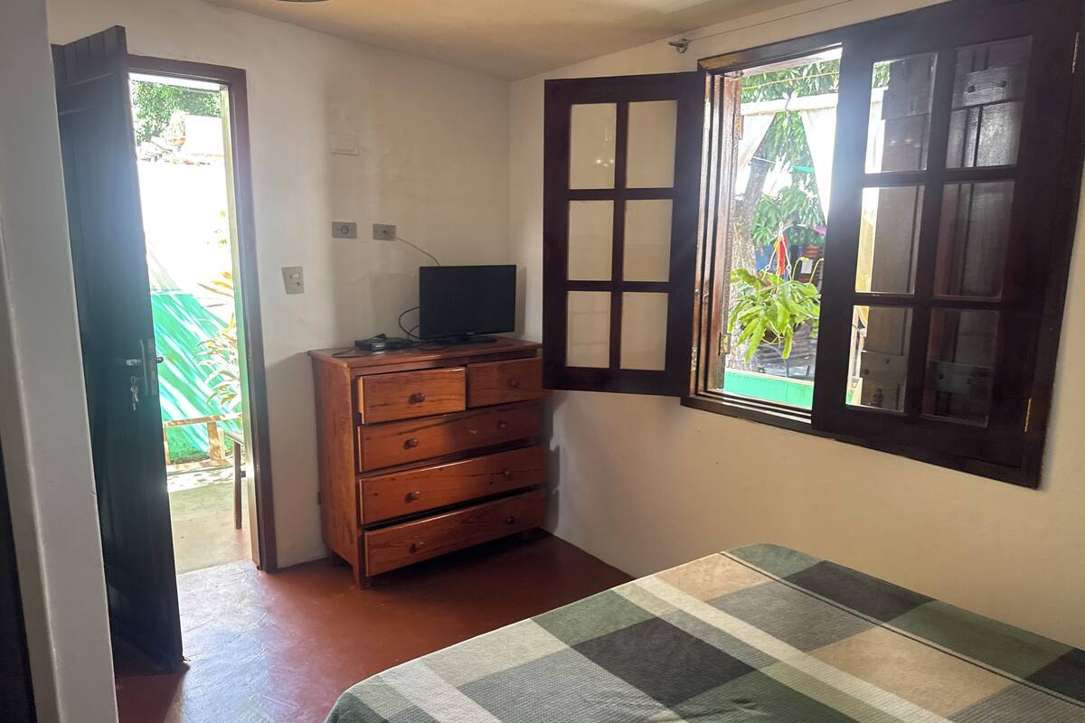 Un dormitorio con cama, cómoda de madera, televisor encima y ventana con vista a follaje.