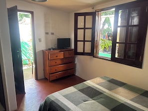 Un dormitorio con cama, cómoda de madera, televisor encima y ventana con vista a follaje.