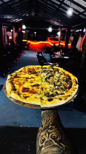 Pizza sobre una tabla de madera sostenida por una persona con un brazo tatuado.